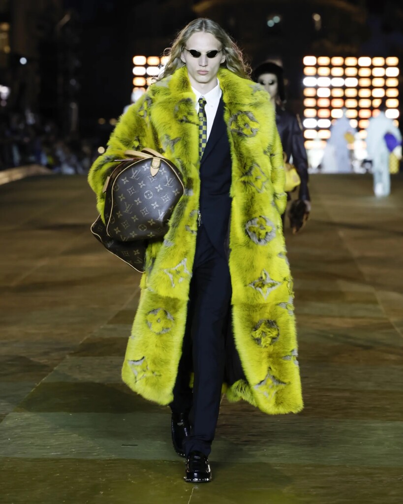 Un mannequin masculine défile à Paris pour Louis Vuitton. Il porteur manteau jaune très long en fourrure. Dans la fourrure, on lit le monogramme LV de Louis Vuitton.