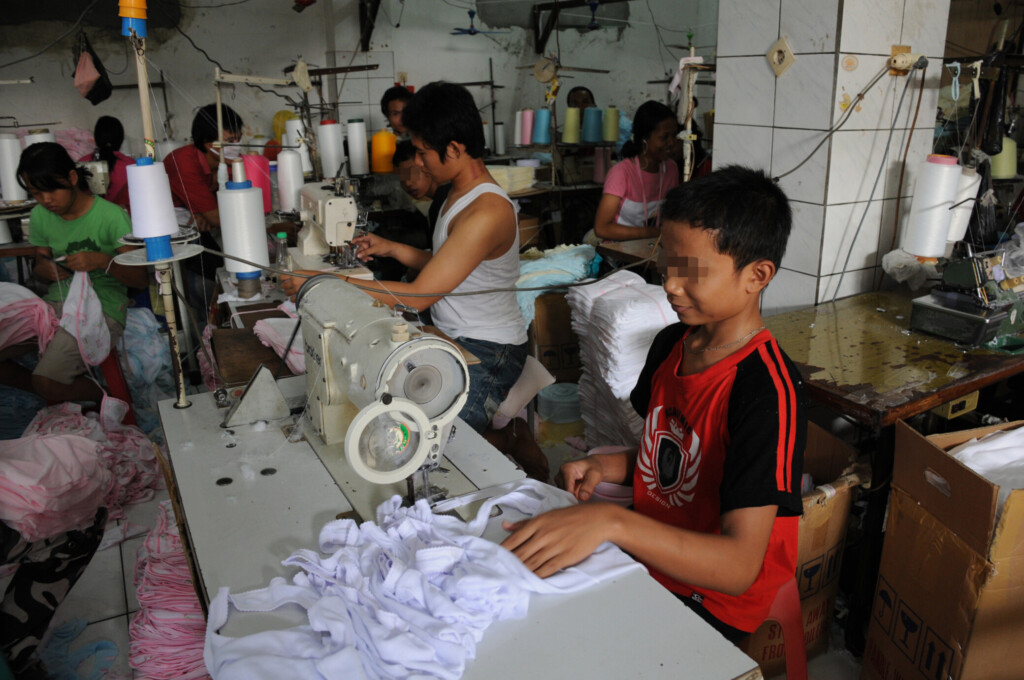 Des enfants travaillent dans une usine textile, d'abouts devant des tables avec des tissus et des machines à coudre.