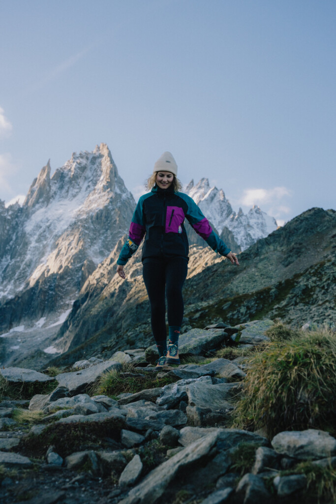 Une mannequin porte une veste, un pantalon outdoor, un bonnet et des baskets issues de la collaboration entre Picture et la Sportiva, dans un decor de montagne